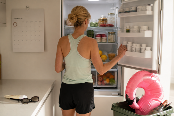 Blonde lady in running clothes looking into healthy fridge. Pink flamingo inflatable and rese wine bottle in recycling bin,